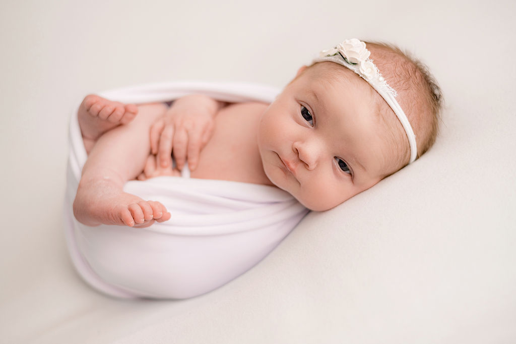 newborn baby wrapped in white and wearing a white headband with her eyes open Pediatricians in Toronto